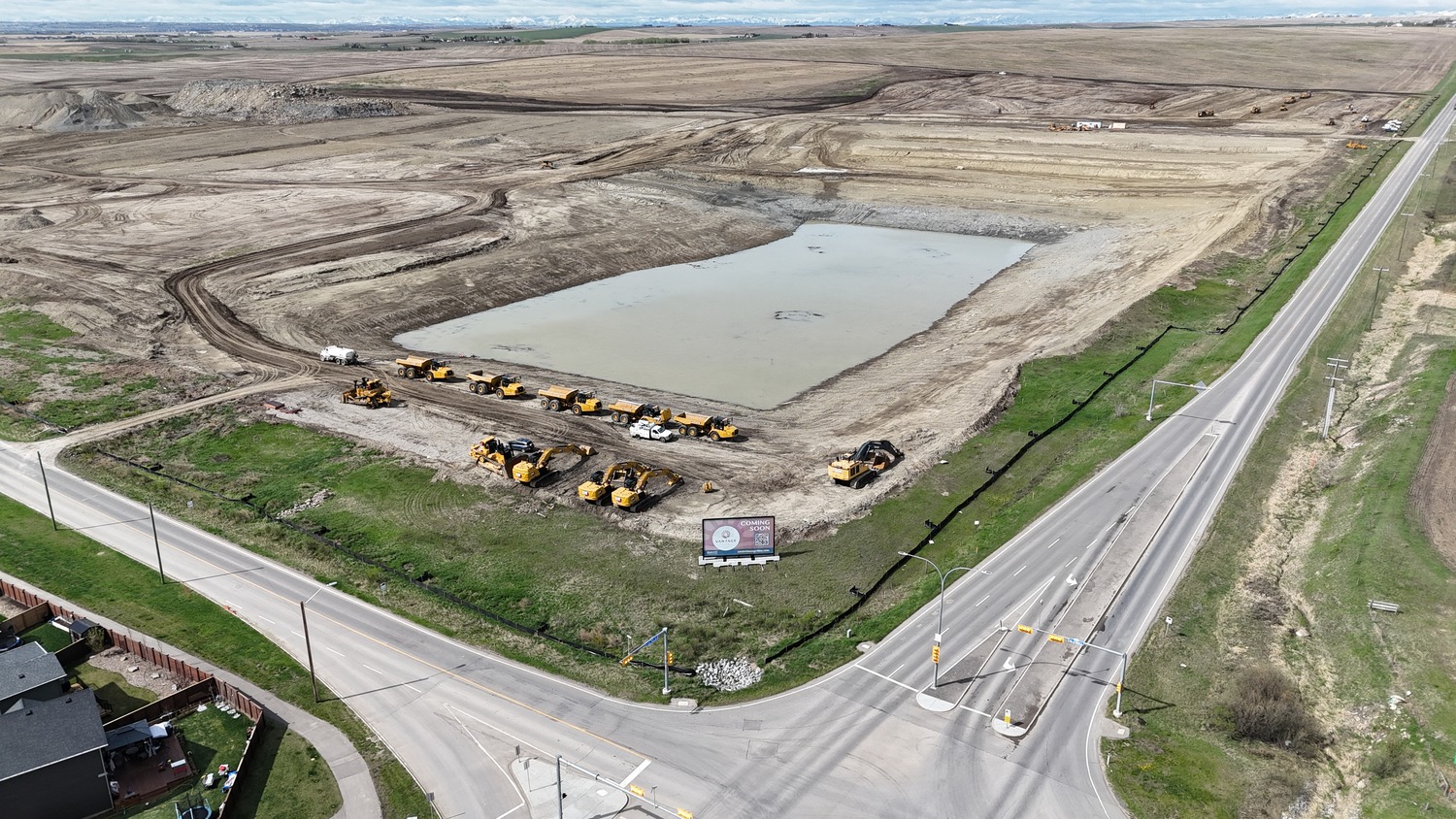Aerial view of a construction site in the Vantage Rise community, with several yellow heavy machinery vehicles parked around a large water-filled excavation next to intersecting roads.
