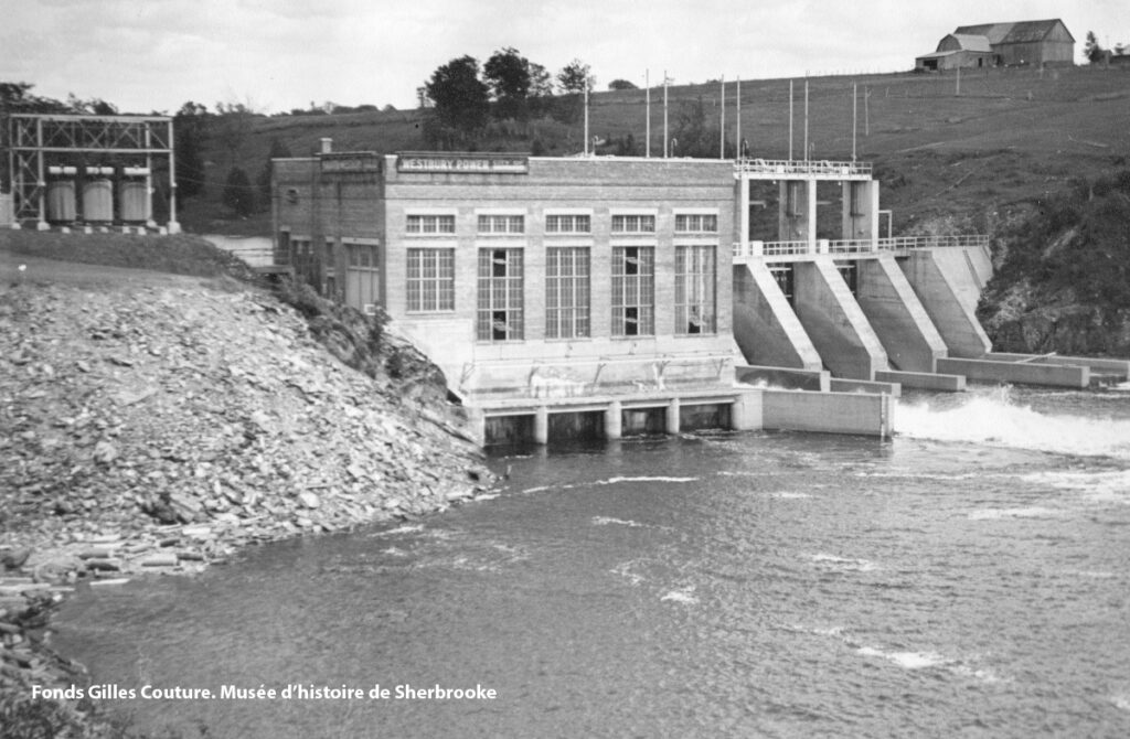 Black and white photo of a hydroelectric power station beside a river, with spillway gates open and water flowing. Hills and a small building are visible in the background.
