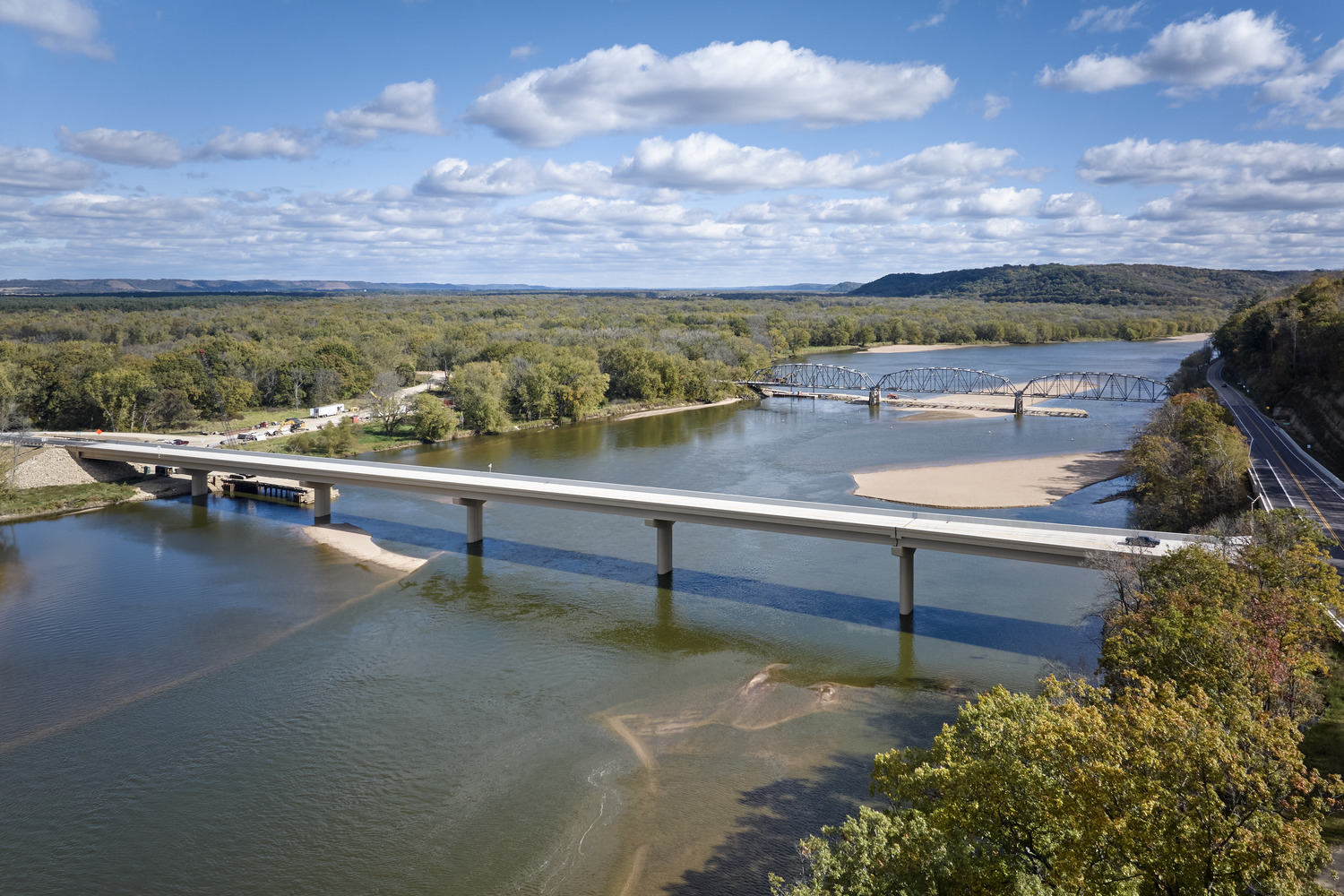 A concrete bridge and an older metal truss bridge span a wide river surrounded by trees under a partly cloudy sky.