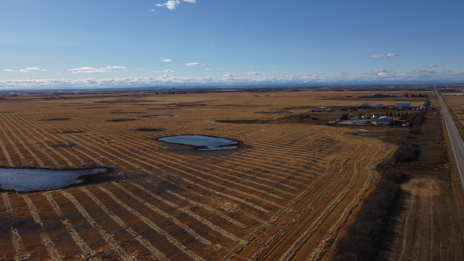 Aerial view of large, flat farmland near the Prairie Economic Gateway with two ponds, striped fields, scattered buildings, and a road running along the right edge under a blue sky.