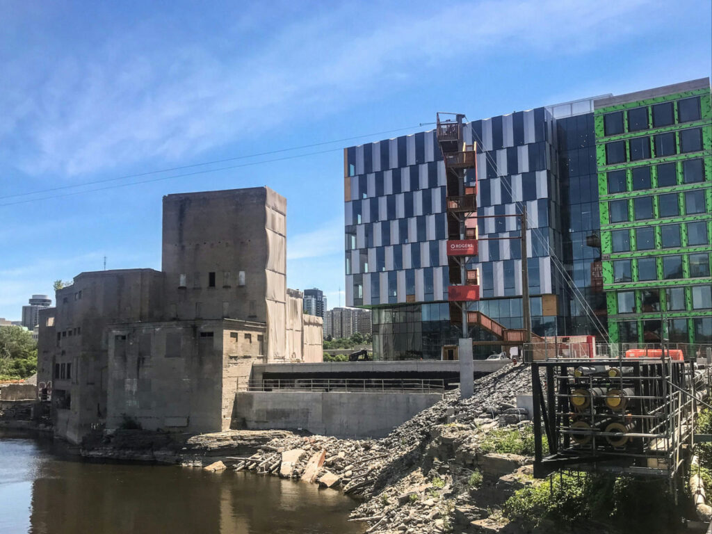 A modern building under construction beside an older structure by a river, amidst a rocky shoreline.