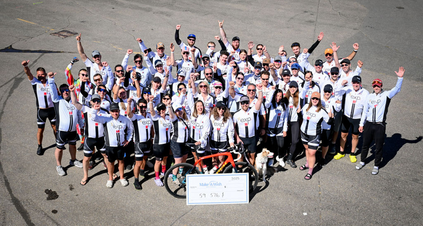 A large group of cyclists wearing matching jerseys poses together outdoors, raising their arms in celebration around a large check and an orange bicycle.