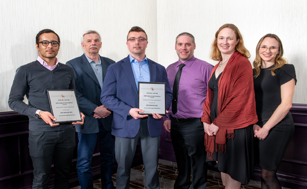 Six people stand indoors, with two men holding framed award plaques for the Florenceville Bridge Rehabilitation. All are dressed in business or semi-formal attire and are facing the camera.