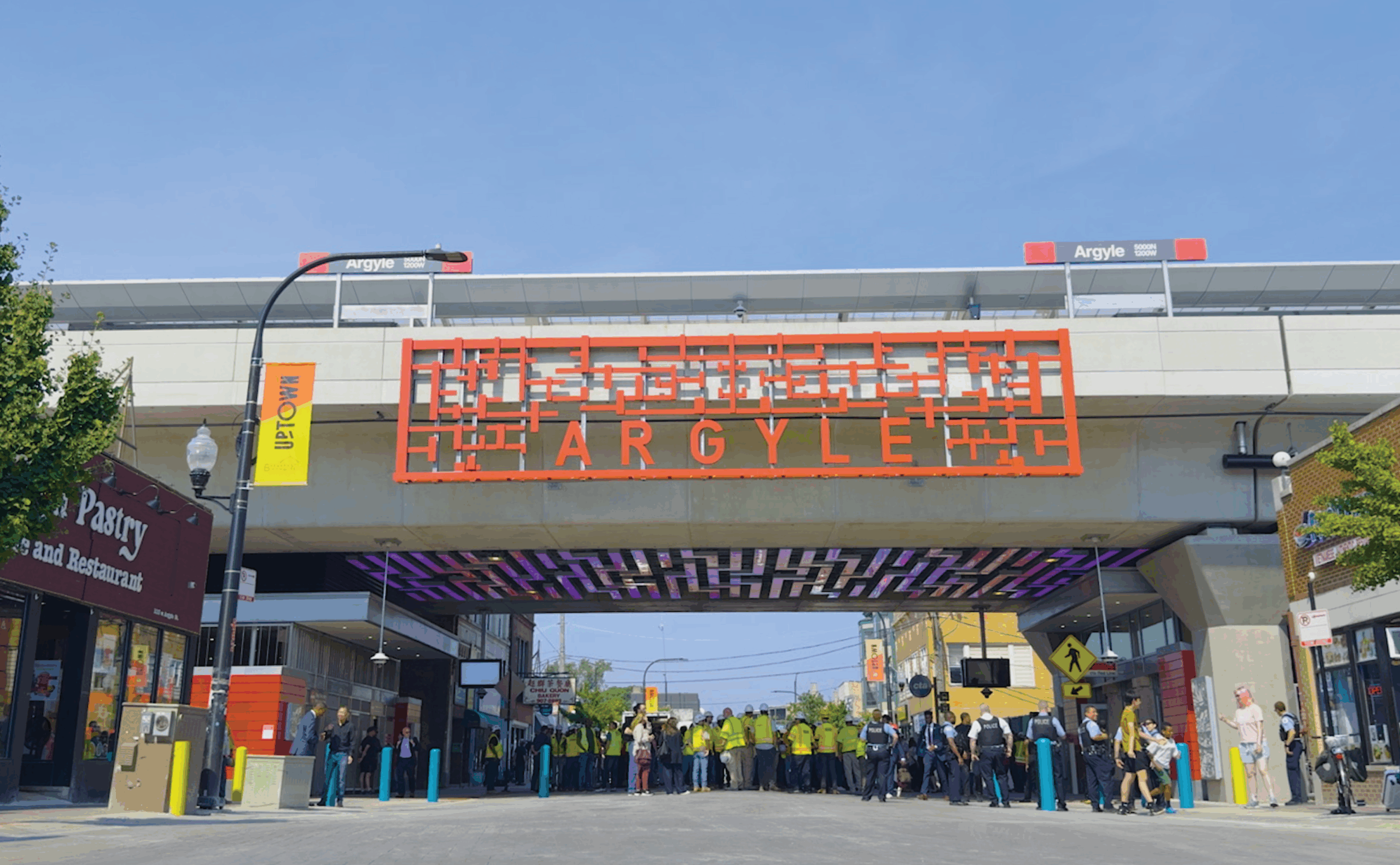 A group of people gathers beneath the Argyle CTA Red Line station with an orange decorative sign, shops on either side, and clear sky above.