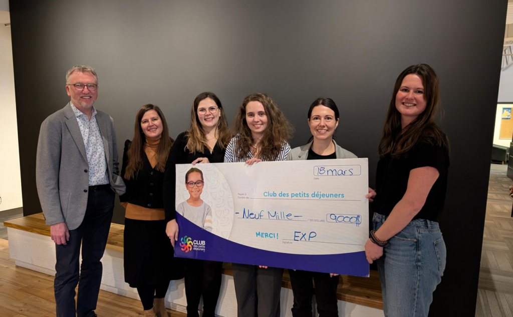 Seven people stand indoors holding a large, novelty check with a logo and signature, dated March 18.