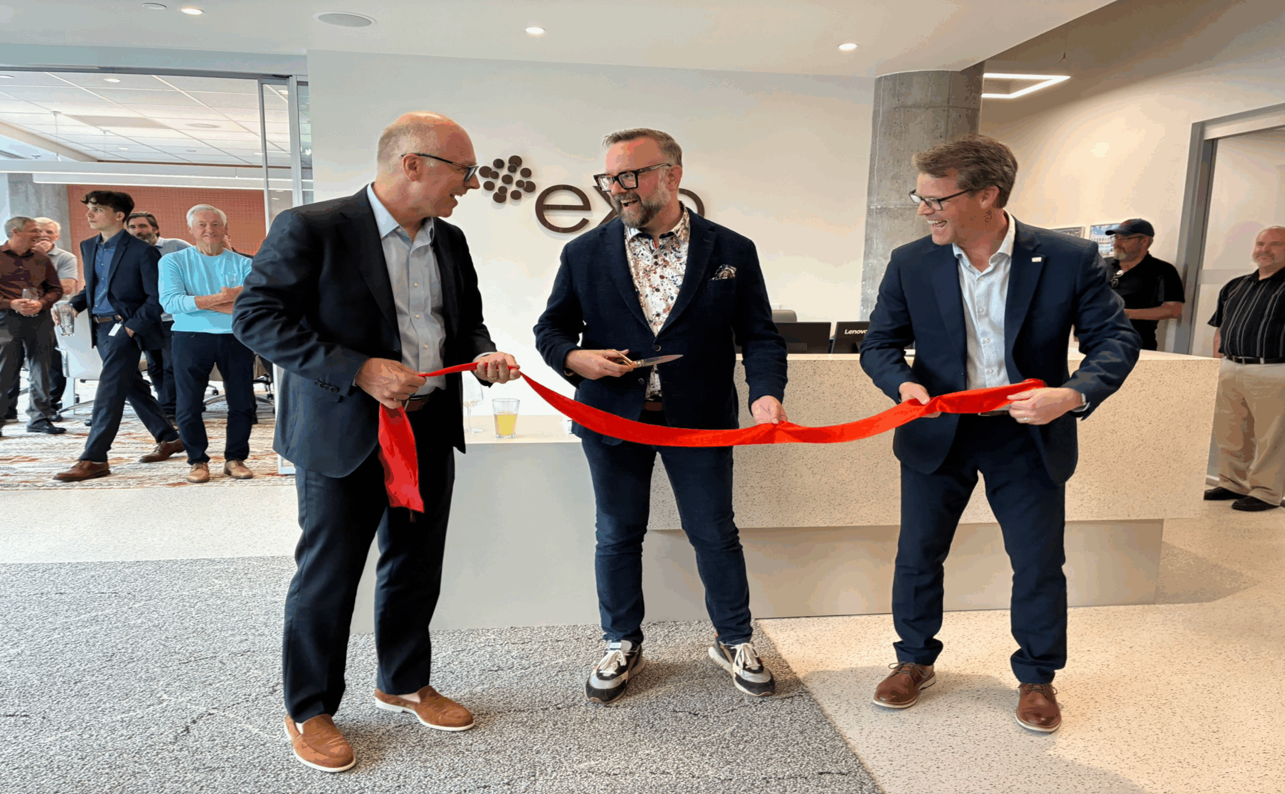 Three men in suits stand together; the man in the center holds scissors, preparing to cut a red ribbon during an indoor ribbon-cutting ceremony with several people watching in the background.