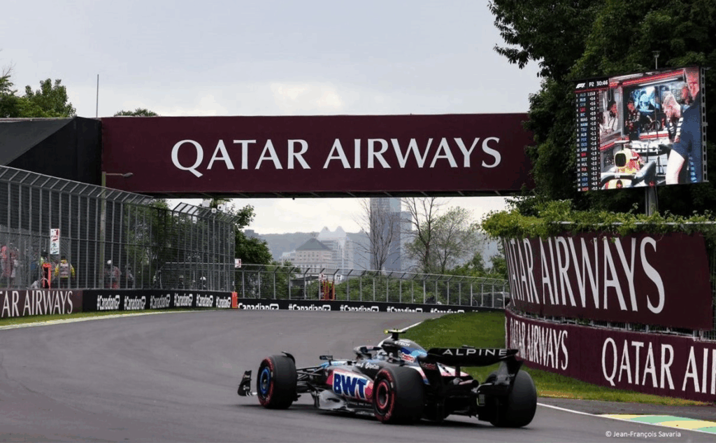 A Formula 1 car drives on a racetrack under a bridge with "Qatar Airways" signage, with trees, a fence, and a distant city skyline in the background.