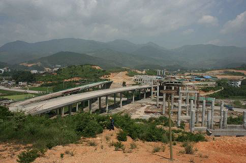 Partially constructed highway overpass and bridge in a rural area with mountains and clouds in the background.