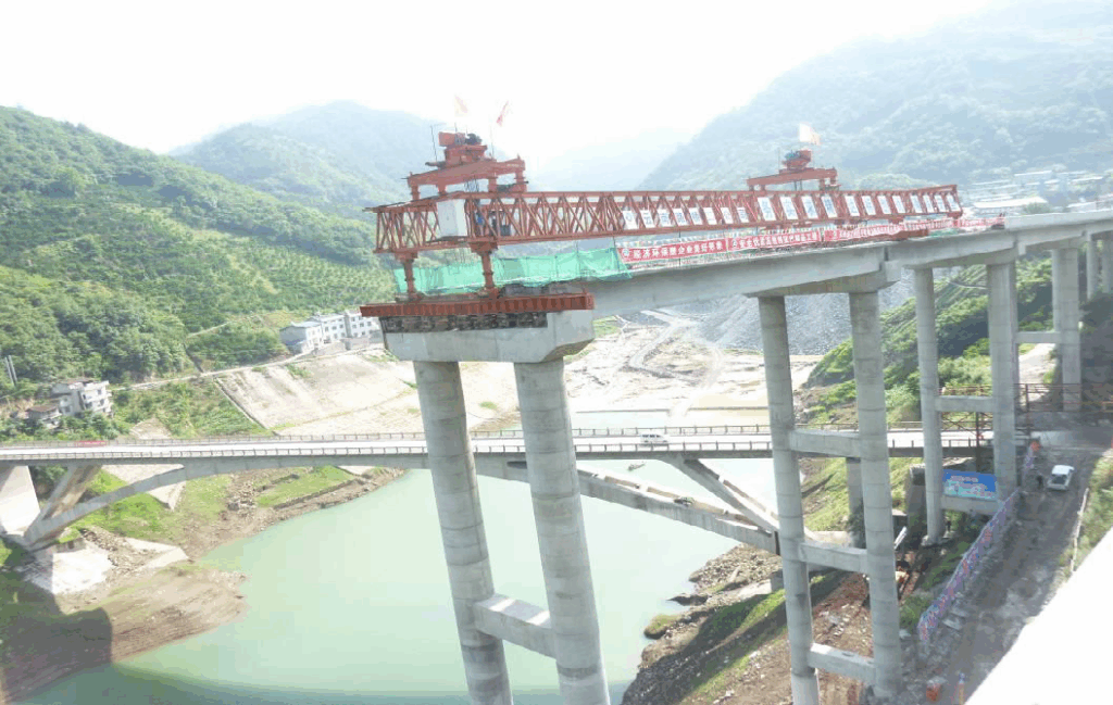 A partially constructed bridge spans over a river in a mountainous area, with scaffolding and construction equipment visible on the bridge structure.