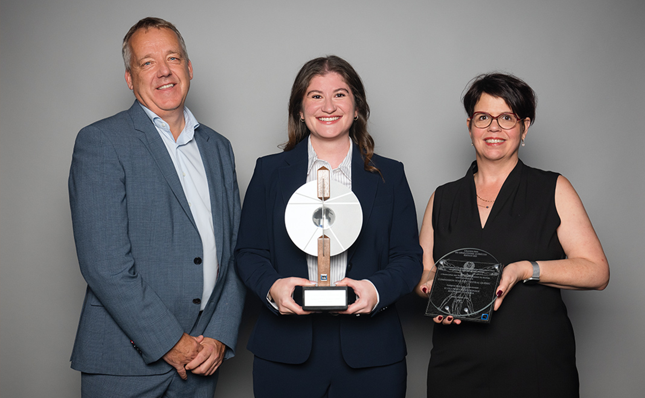 Three people in formal attire stand against a plain background; the center person holds a large trophy, and the person on the right holds a glass award.