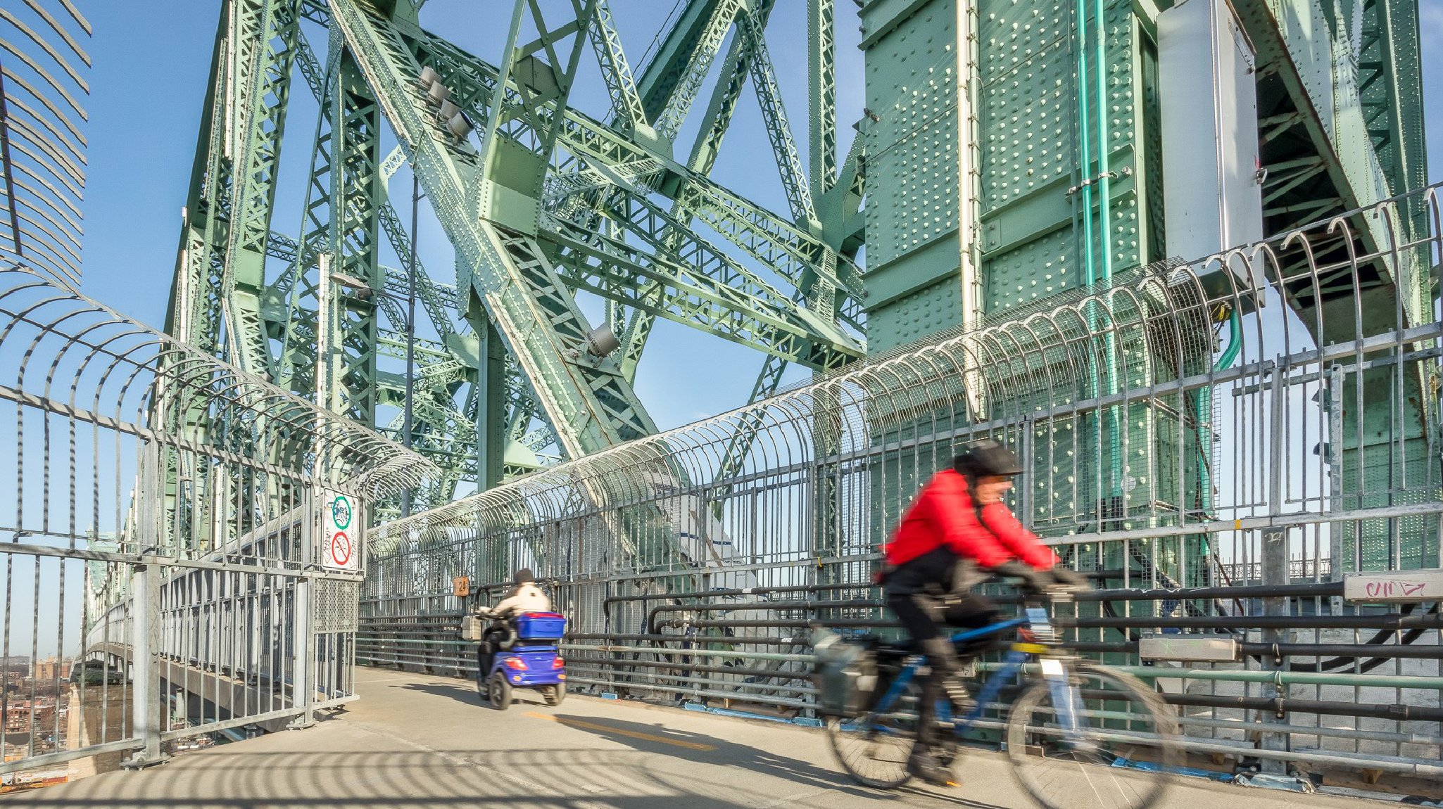 A cyclist and a person in a mobility scooter travel on a fenced bridge pathway with large green steel beams in the background.