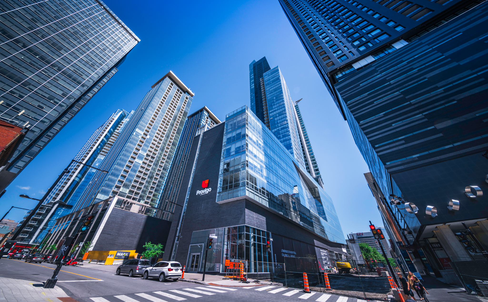 Tall modern skyscrapers surround a city intersection with crosswalks, construction barriers, and a building displaying the "Paddyp" logo under a clear blue sky.