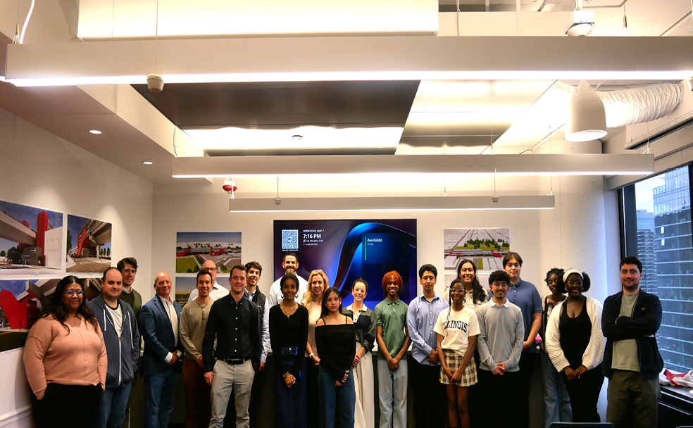 A group of people stand together in an office meeting room at Lindblom Math and Science Academy, posing for a group photo in front of presentation screens and architectural posters.