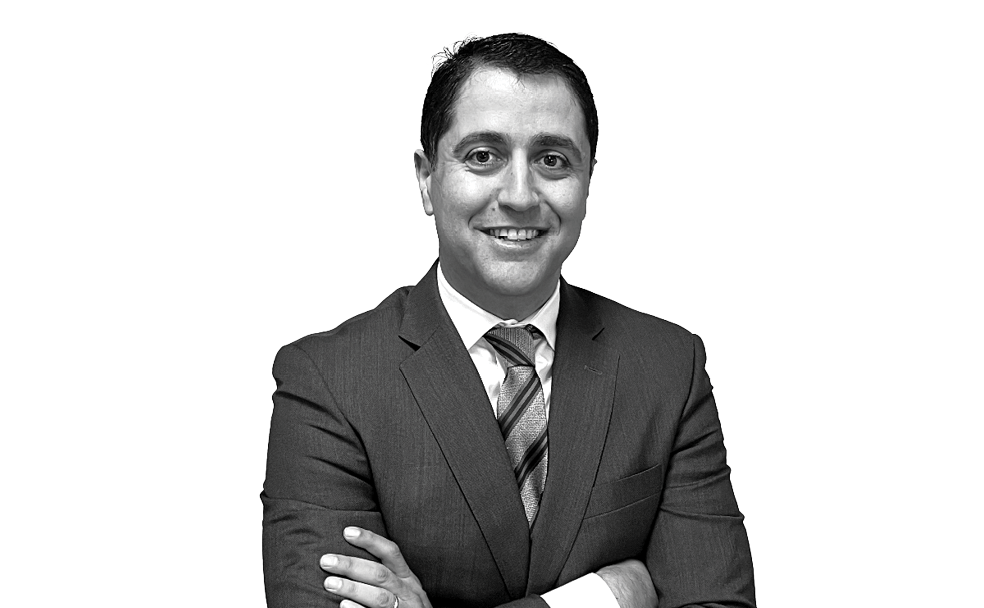 A person wearing a suit and tie is smiling at the camera with their arms crossed in a studio setting against a white background.