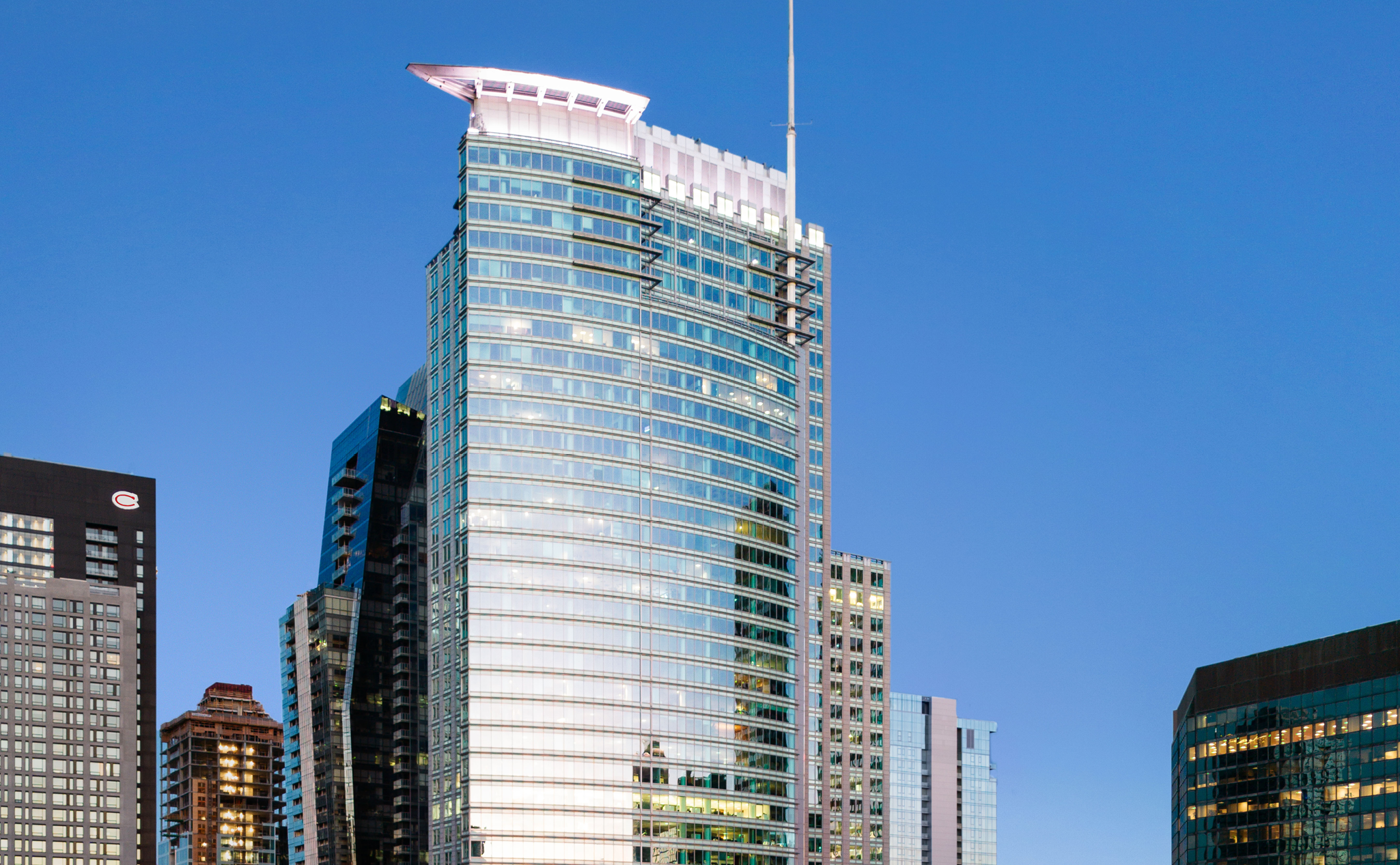 A modern glass skyscraper with a curved facade and unique white rooftop feature, reflecting surrounding buildings under a clear blue sky.