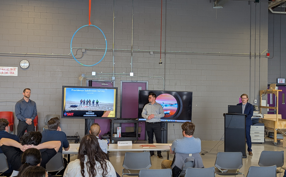 Three people from SAIT Connector Lab stand at the front of a room giving a presentation to an audience, with two large screens displaying slides behind them.
