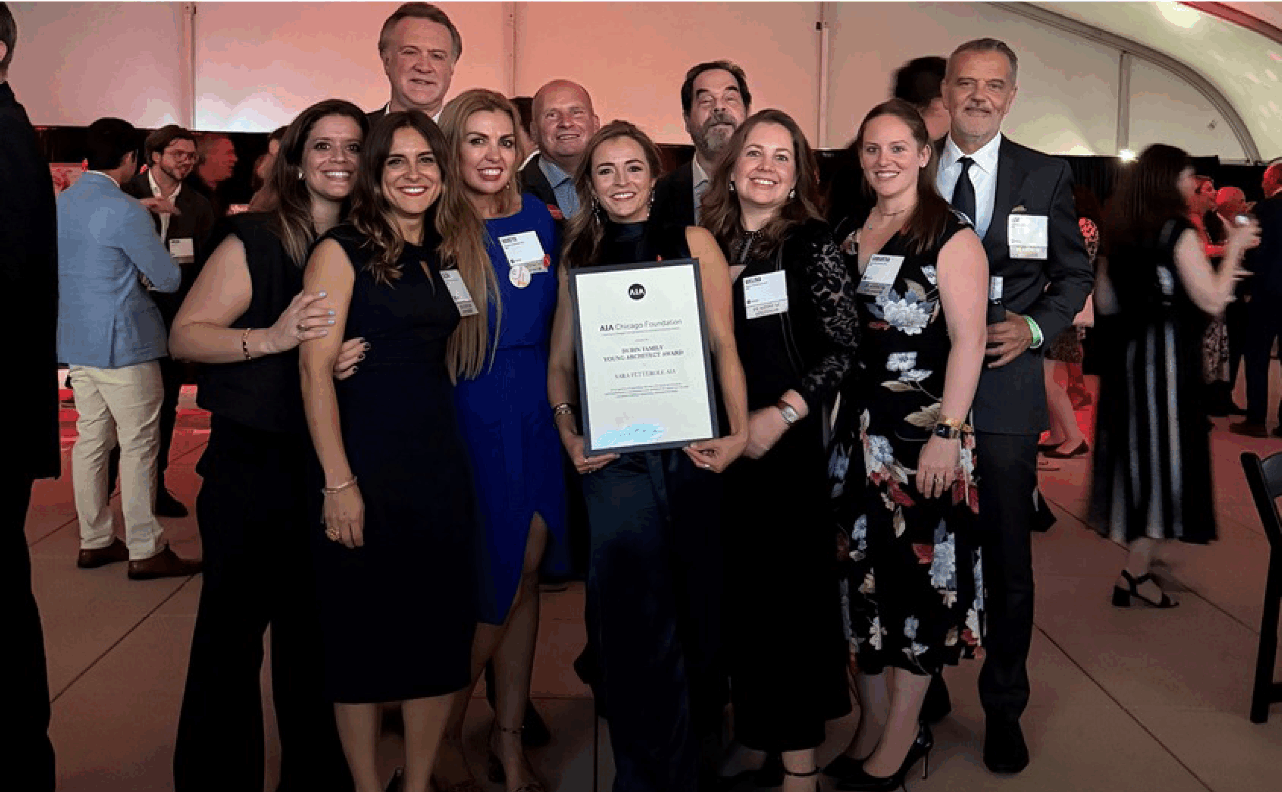 A group of people dressed in formal attire pose together indoors, with one person in the center holding a large framed certificate.